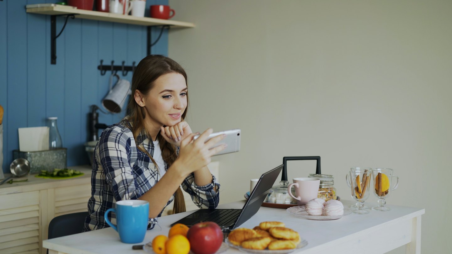 Woman smiling at phone on kitchen table with breakfast.